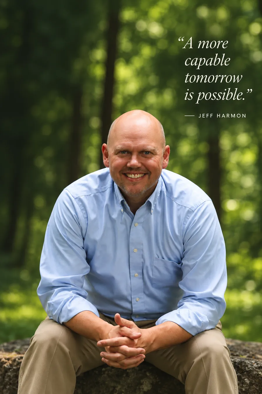 Jeff D. Harmon seated on a stone in a sunlit forest, hands clasped, smiling